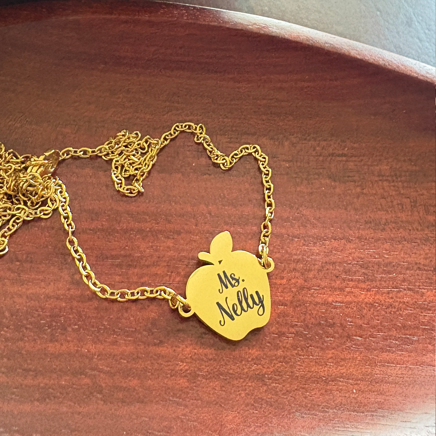 Close up of a gold tone stainless steel apple charm necklace with engraved words that says Ms. Nelly