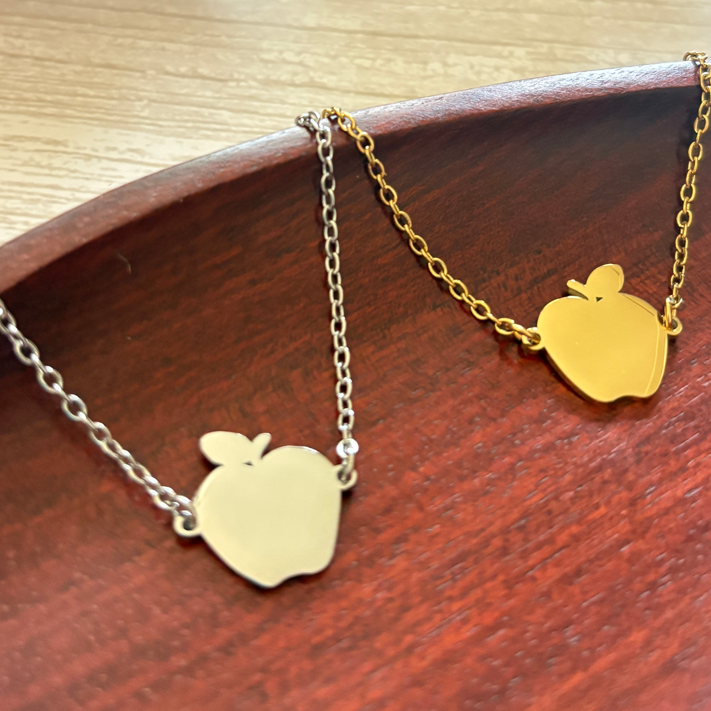 2 Apple-shaped stainless steel necklace one in silver and one in gold finish, displayed on a wooden tray