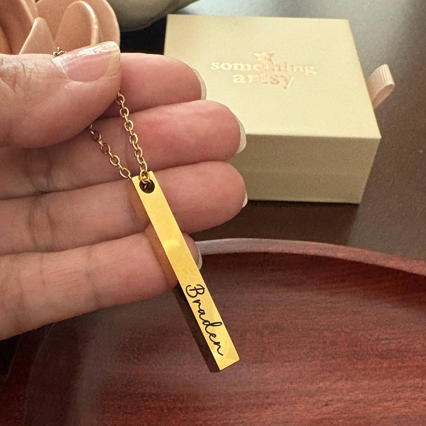 Gold necklace with a bar pendant engraved with the name 'Braden' held by a hand, with a 'something artsy' box in the background.