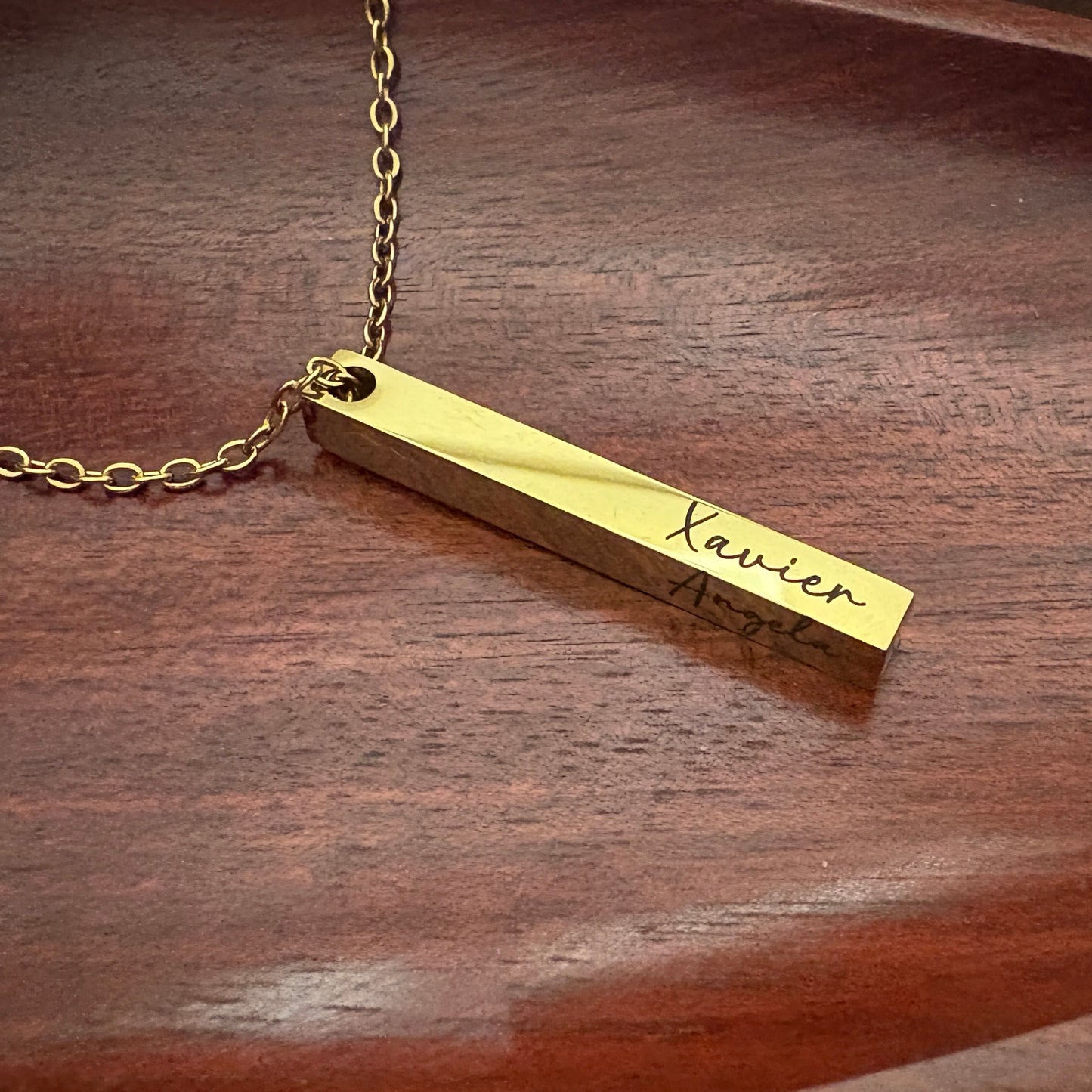 Gold necklace with a bar pendant on a wooden surface showing the names engraved in both sides.