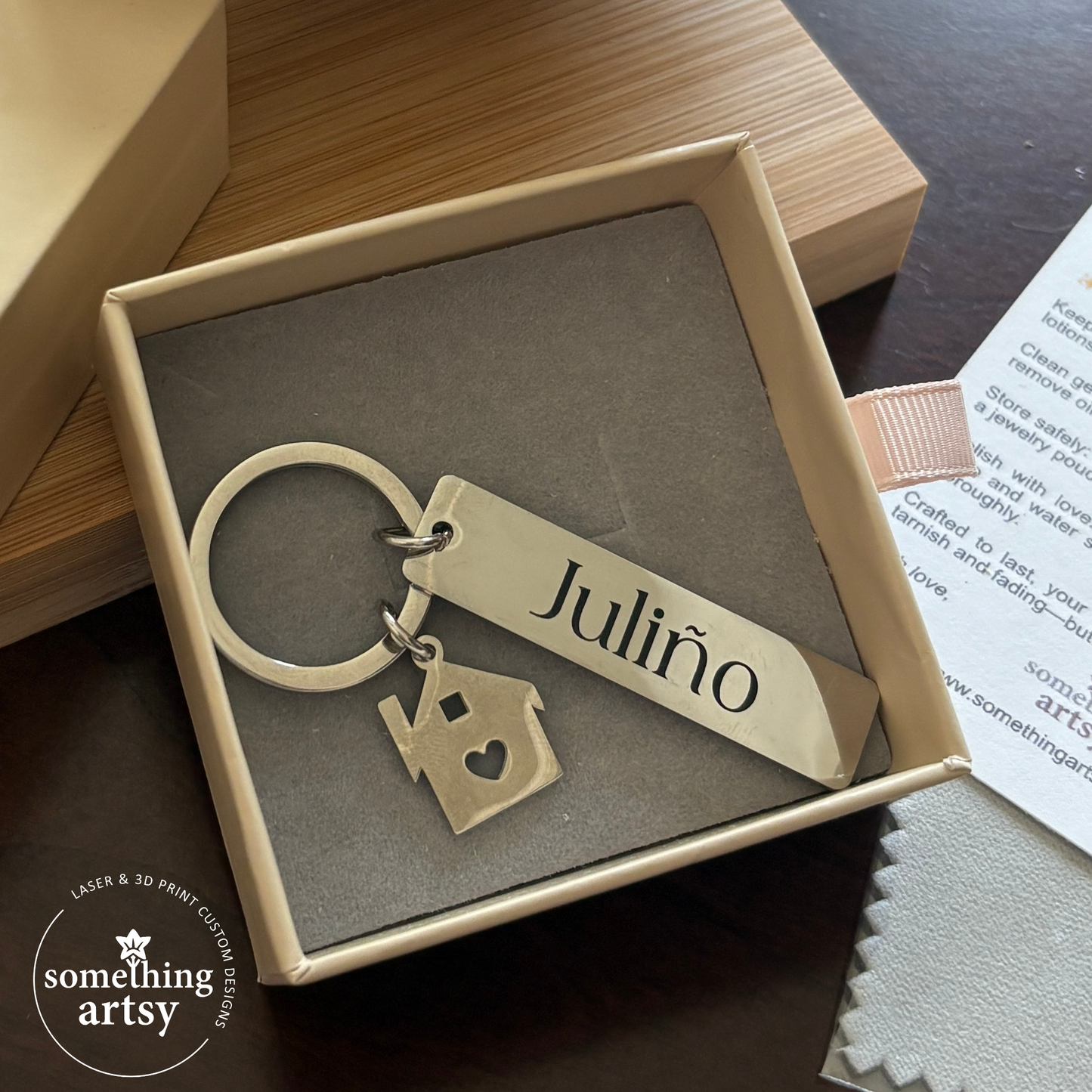 Stainless steel keychain with a house charm and a rectangle engraved with a name inside a package box
