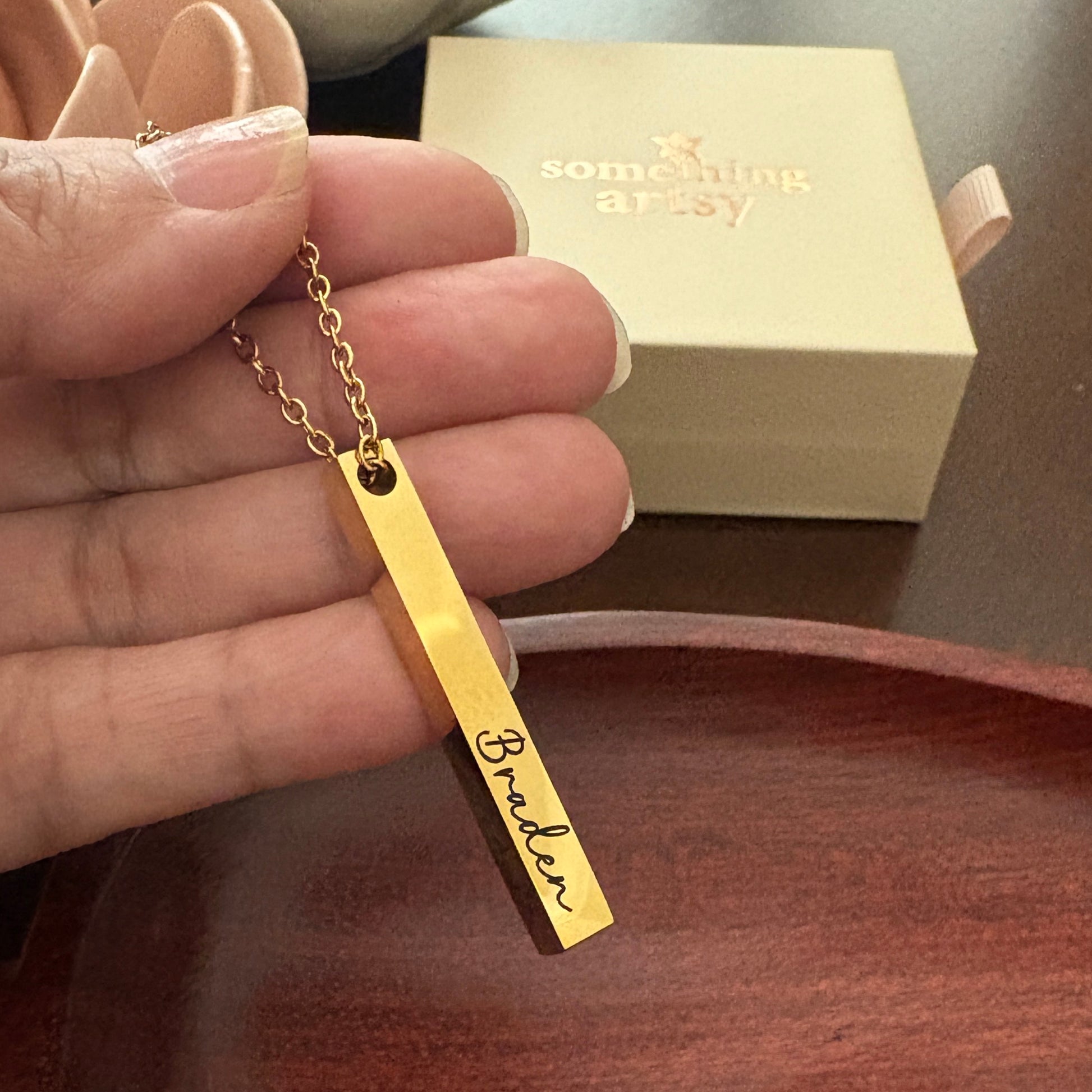 Gold necklace with a bar pendant engraved with the name 'Braden' held by a hand, with a 'something artsy' box in the background.