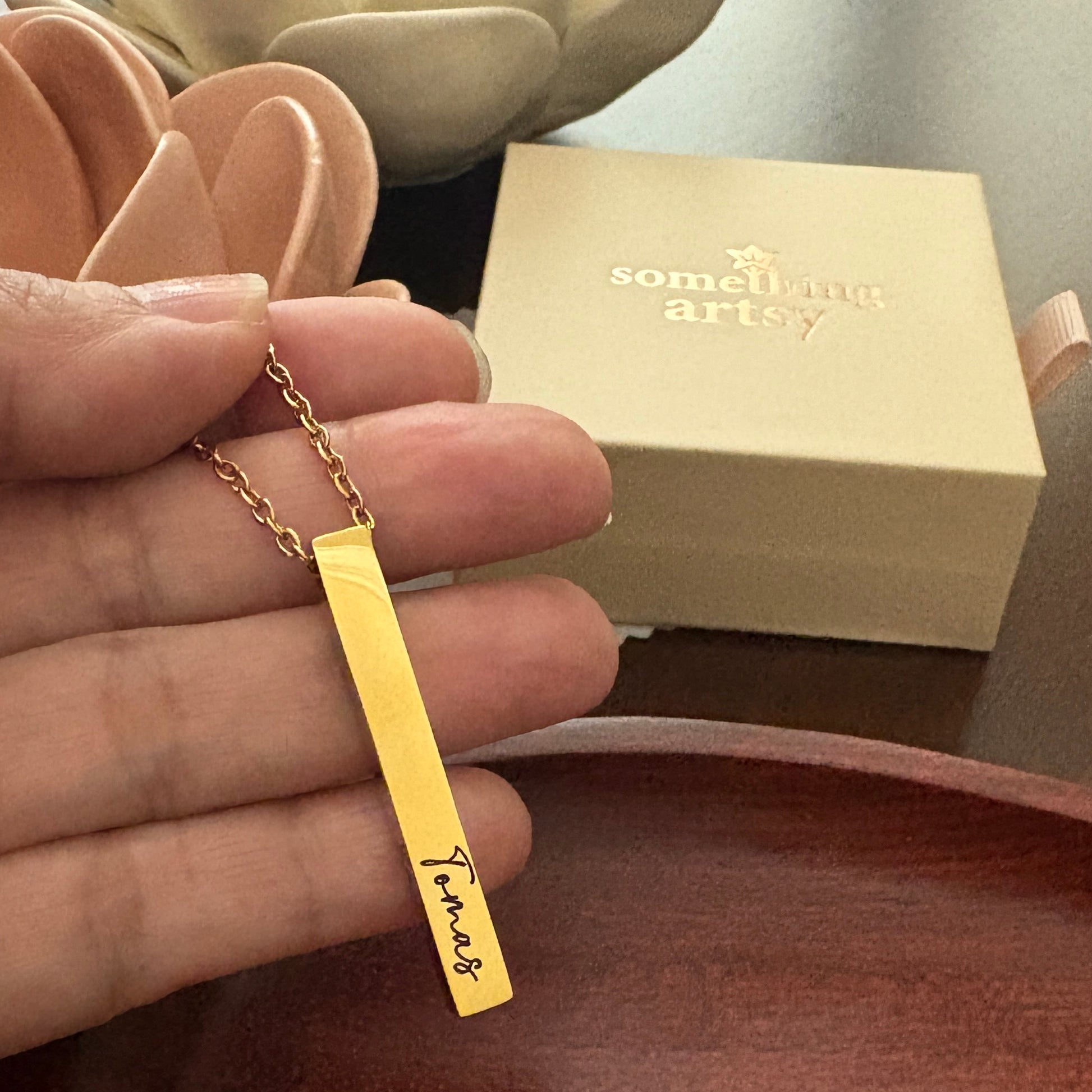 Gold necklace with a bar pendant engraved with the name "Tomas" held by a hand, with 'something artsy' box in the background.