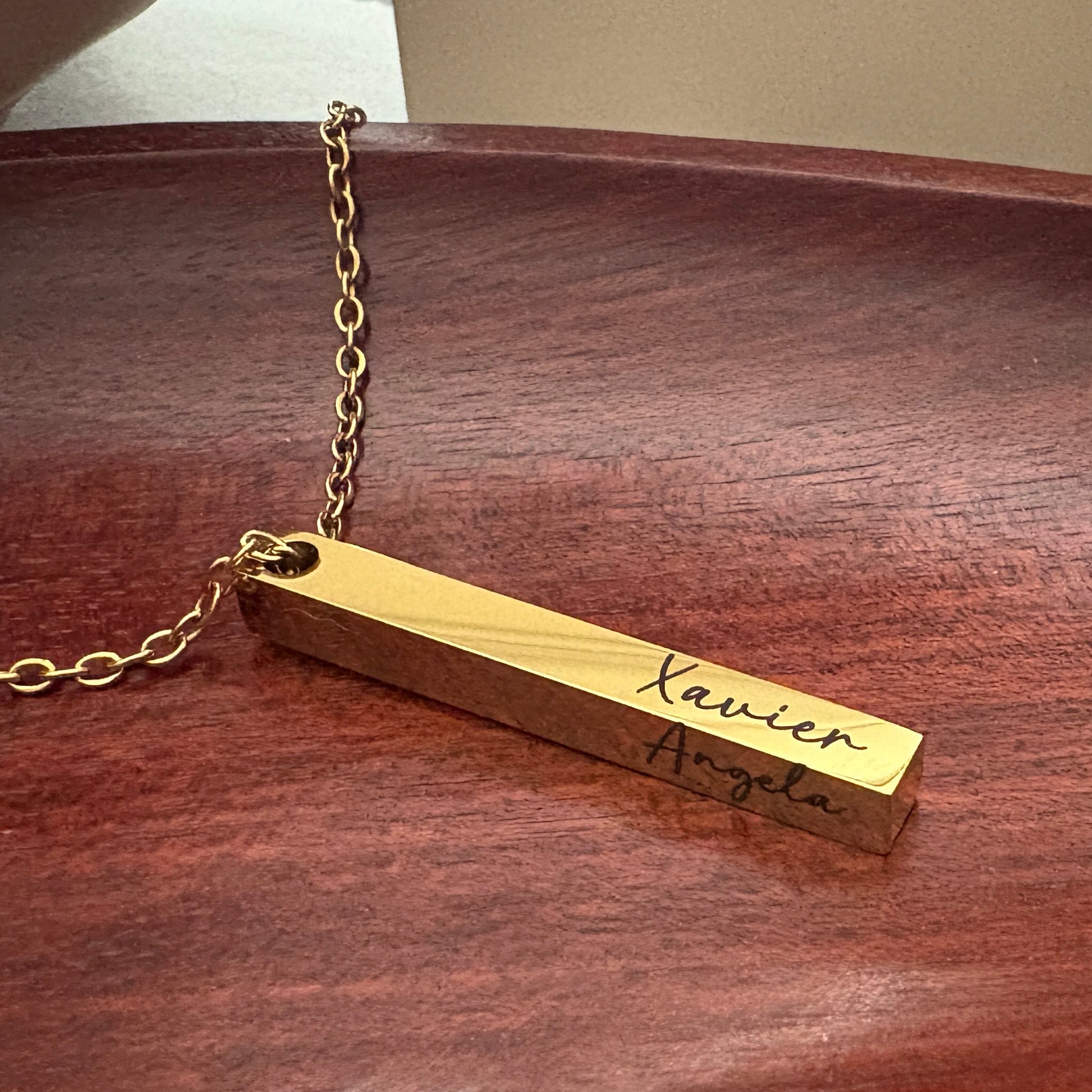 Gold necklace with a bar pendant showing the engraved names  'Xavier and Angela' on both sides.