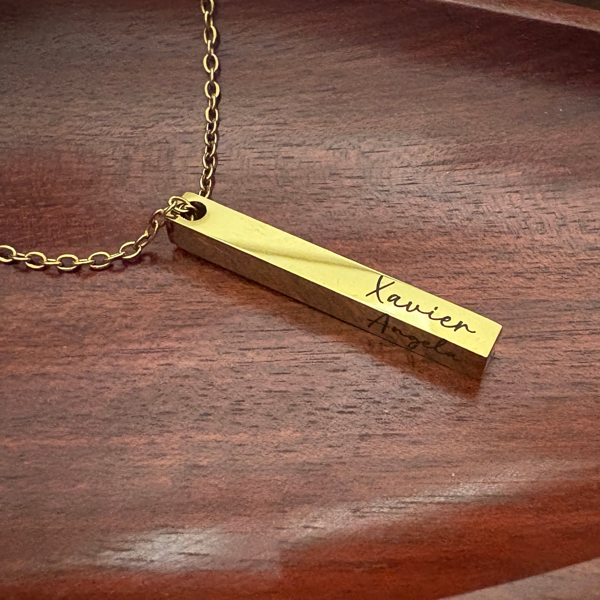 Gold necklace with a bar pendant on a wooden surface showing the names engraved in both sides.