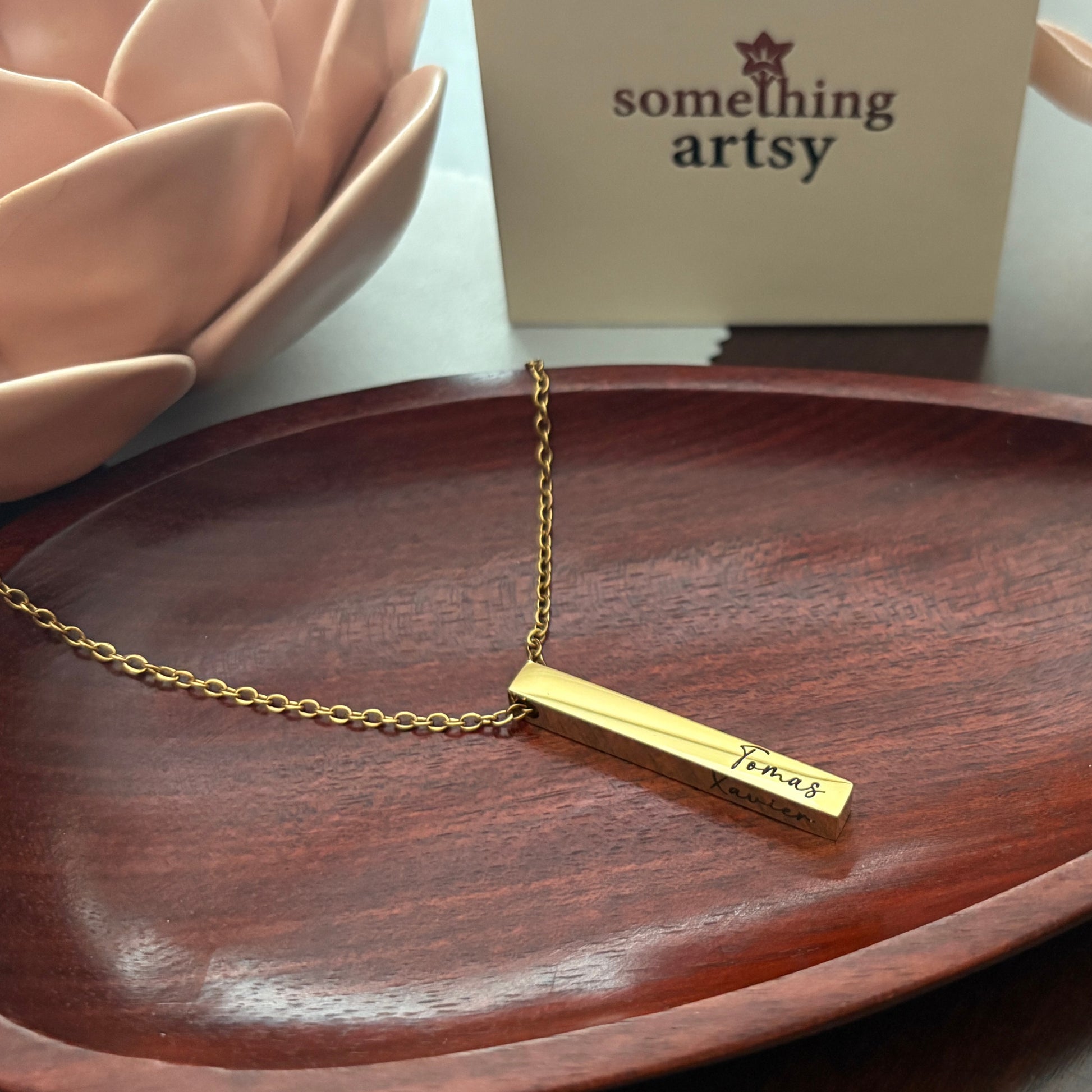 Gold necklace with a bar pendant engraved on a wooden surface, with 'something artsy' box in the background.