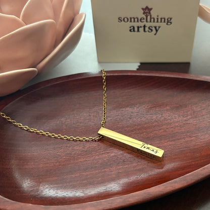 Gold necklace with a bar pendant engraved on a wooden surface, with 'something artsy' box in the background.