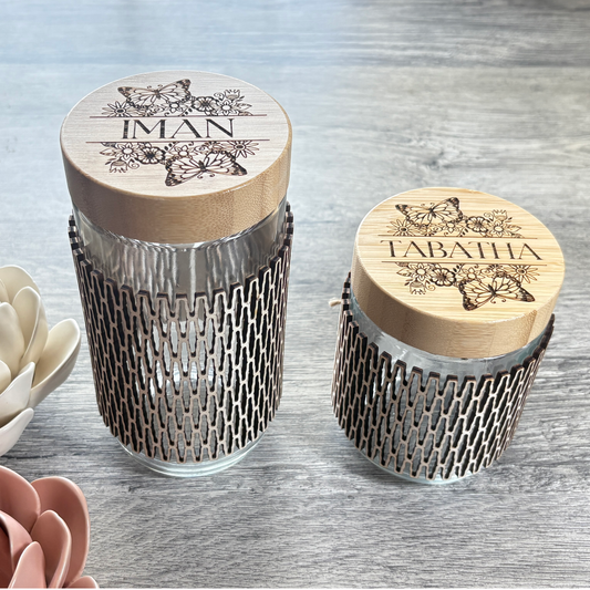 A 24oz glass jar and a 16oz jar side by side both have a wooden sleeve and custom engraving on the bamboo lid.